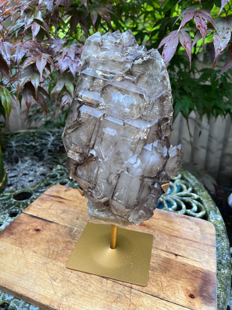 May include: A large, smoky quartz crystal cluster mounted on a gold-colored metal stand. The crystal has a complex, geometric structure with multiple facets and a translucent appearance. The stand has a square base and a slender, vertical support.