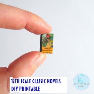 May include: A tiny book, "Anne of Green Gables", is held by fingers. The cover shows a woman in a yellow dress and red hat. The text "12th Scale Classic Novels DIY Printable" is visible. The book is a miniature item.