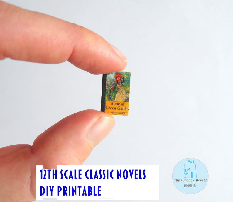 May include: A tiny book, "Anne of Green Gables", is held by fingers. The cover shows a woman in a yellow dress and red hat. The text "12th Scale Classic Novels DIY Printable" is visible. The book is a miniature item.