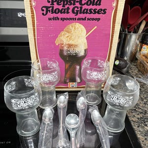 May include: A set of four clear glass Pepsi-Cola float glasses with white logo detailing, accompanied by four metal spoons and one metal ice cream scoop. The items are displayed in front of their original cardboard box featuring pink graphics.