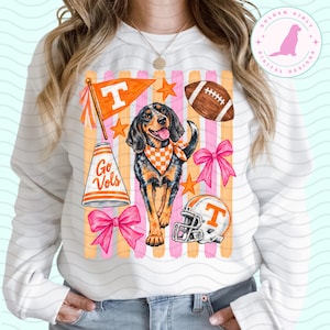 May include: A white long-sleeve sweatshirt featuring a graphic print of a hound dog wearing an orange and white checkered bandana. The design includes an orange pennant with a T, a football, a megaphone with Go Vols, a helmet, and pink bows.