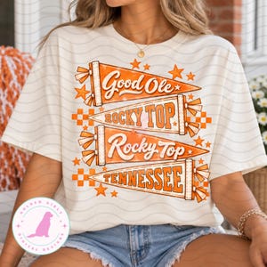 Peut inclure: Un t-shirt couleur crème arborant un motif de quatre fanions orange empilés verticalement. Les fanions affichent le texte Good Ole, ROCKY TOP, Rocky Top et TENNESSEE en orange et blanc, agrémenté d'étoiles orange et de motifs en damier.