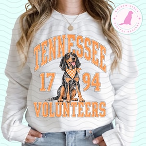 May include: A white long-sleeve sweatshirt featuring a graphic of a black and tan hound dog wearing an orange and white checkered bandana. The text reads TENNESSEE 17 94 VOLUNTEERS in distressed orange block lettering surrounding the dog illustration.