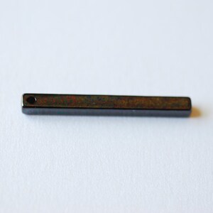 Peut inclure: Un pendentif rectangulaire brun foncé avec un petit trou à une extrémité. Le pendentif a une surface légèrement texturée et est posé sur un fond blanc. Le pendentif mesure environ 5 cm de long.