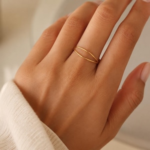 May include: A thin gold ring featuring a double-band design that creates an open, elongated oval shape across the finger. The ring is crafted from a smooth, polished gold-toned metal, showcasing a delicate and minimalist structure on the finger.