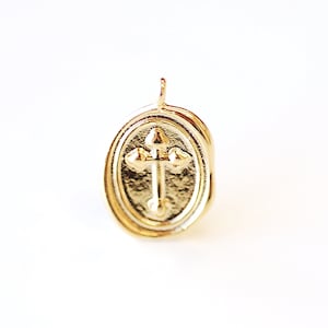 Puede incluir: Un colgante ovalado de oro con un diseño de cruz. El colgante tiene un borde elevado y una superficie lisa.