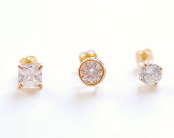May include: Three gold stud earrings with clear cubic zirconia stones. The earrings feature a square, round, and round stone.
