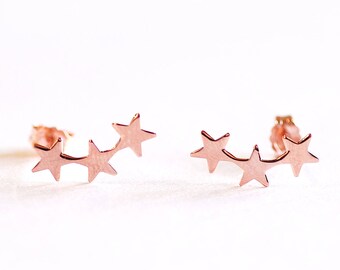 May include: A pair of rose gold star earrings. Each earring features three small stars connected together in a curved shape.