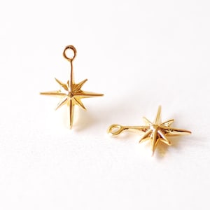 Pode incluir: Dois pingentes dourados em forma de estrela com um laço no topo. Os pingentes têm um design de estrela pontiaguda com oito pontas.