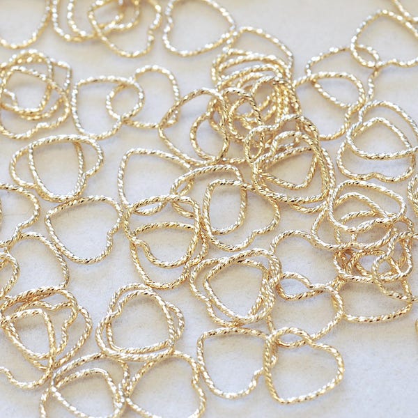 Puede incluir: Una colección de pequeños dijes de metal de color dorado con forma de corazón. Cada dije presenta un diseño de alambre trenzado y texturizado que forma un contorno de corazón abierto y hueco. Los dijes están dispersos en un arreglo suelto.