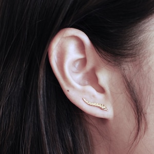 Puede incluir: Primer plano de la oreja de una persona con un pendiente de hoja de oro. El pendiente es una pieza de joyería delicada que añade un toque de elegancia a cualquier atuendo.