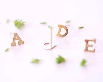Puede incluir: Pendientes de letras doradas con las letras A, D y E. Los pendientes están sobre un fondo blanco.