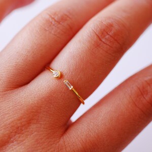 Può includere: Un anello d'oro con due piccole pietre trasparenti. L'anello è aperto da un lato e ha un design delicato.