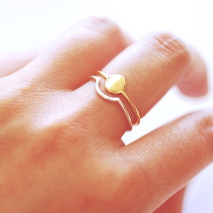 May include: A gold ring with a round, flat top and a thin band. The ring is worn on the finger of a hand.