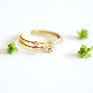 May include: A set of three delicate gold rings, each adorned with a small, sparkling diamond. The rings are arranged in a layered style, creating a modern and elegant look. Small green succulents are scattered around the rings on a white background.