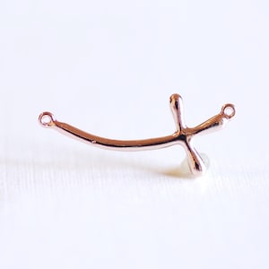 May include: A rose gold-toned metal cross charm with two jump rings. The cross is stylized with a curved, organic shape.