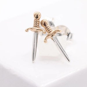 May include: A pair of silver dagger earrings with gold-coloured detailing. The earrings are presented on a white surface. The dagger design features a detailed hilt and a pointed blade, a unique and stylish accessory.