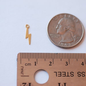 May include: Gold-toned lightning bolt charm pendant with a loop at the top. The charm is about 1 inch tall and is shown next to a US quarter for size comparison.