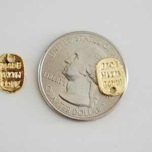 May include: Two gold-coloured metal tags with text. The tag on the left says "Made with Love". The tag on the right says "Love Pizza Now". The tags are shown next to a US quarter.