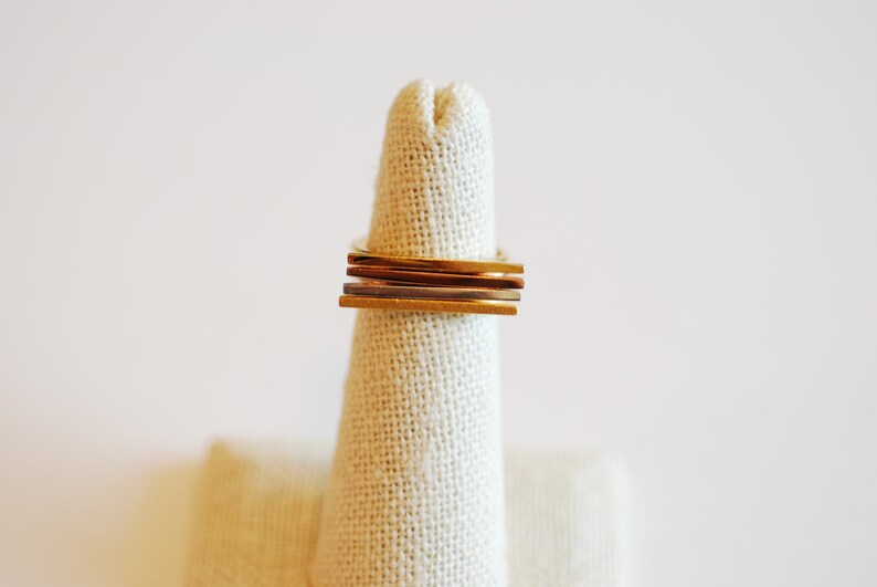 May include: A stack of four gold, silver, and copper rings on a fabric ring display stand.