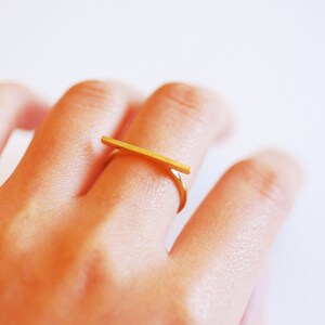 May include: A gold bar ring with a simple, minimalist design. The ring is worn on the index finger of a hand.