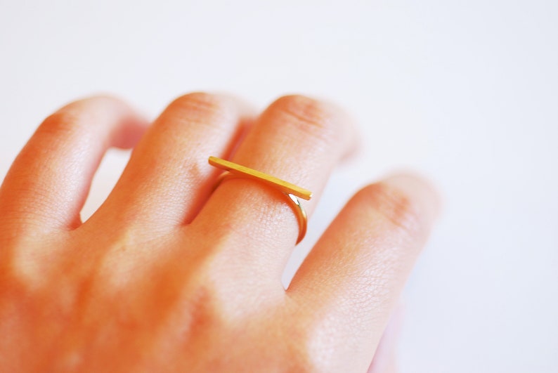 May include: A gold bar ring with a simple, minimalist design. The ring is worn on the index finger of a hand.
