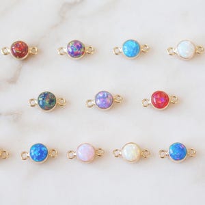 Puede incluir: Una colección de catorce pequeños conectores de joyería redondos estilo cabujón con engastes de metal dorado. Cada pieza presenta una piedra de ópalo sintético lisa y colorida en varios tonos, incluyendo rojo, azul, morado, blanco y rosa.