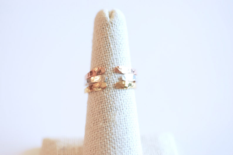 May include: A set of three hammered metal rings in gold, silver, and rose gold tones. The rings are stacked on a fabric ring holder.