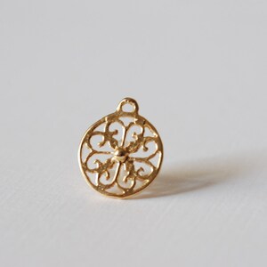 May include: Gold-toned metal charm with a circular, intricate, filigree design. The charm has a loop at the top for attaching to a necklace or bracelet.