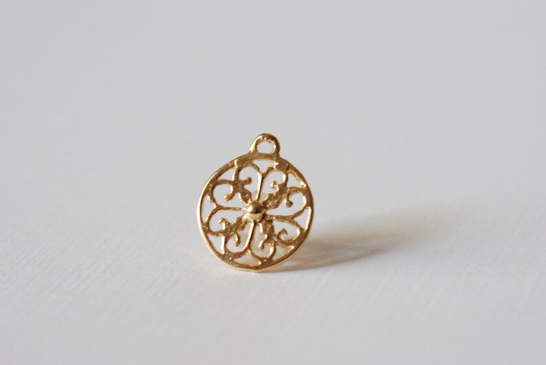 May include: Gold-toned metal charm with a circular, intricate, filigree design. The charm has a loop at the top for attaching to a necklace or bracelet.