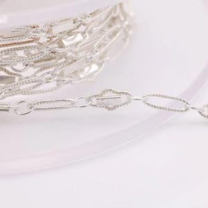 May include: Silver chain with a delicate, textured design. The chain is coiled on a white surface and a section of the chain is extended to show the detail.