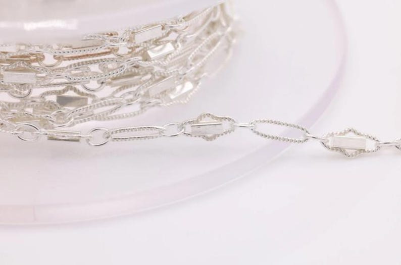 May include: Silver chain with a delicate, textured design. The chain is coiled on a white surface and a section of the chain is extended to show the detail.