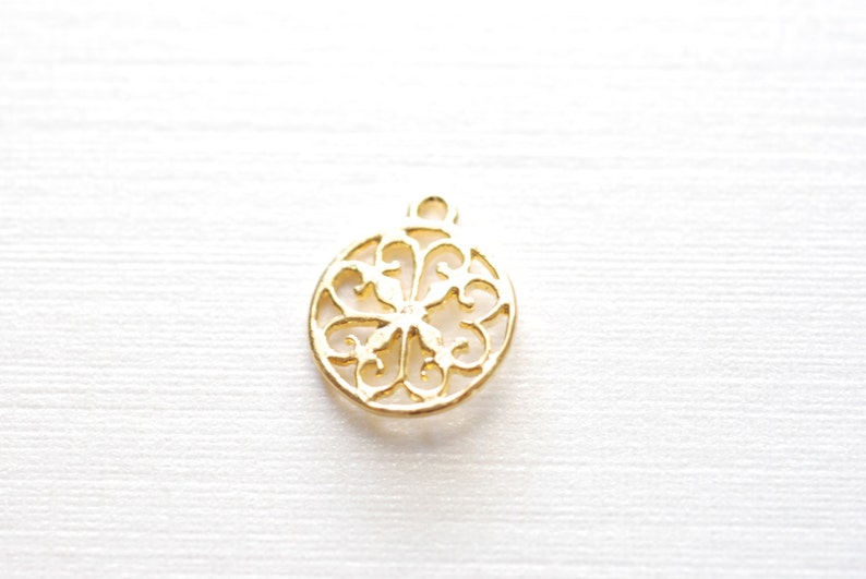 May include: Gold-toned metal ornament charm with a delicate, intricate design. The charm is round and features a cutout pattern of swirls and loops.