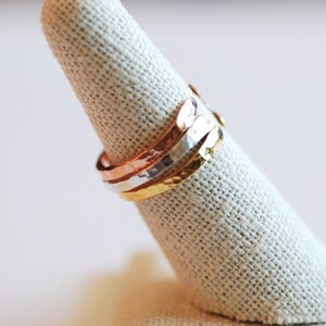 May include: Three hammered metal rings stacked on a fabric ring holder. The rings are gold, silver, and copper.