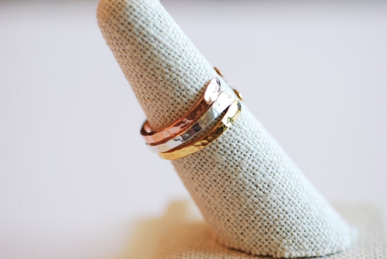 May include: Three hammered metal rings stacked on a fabric ring holder. The rings are gold, silver, and copper.
