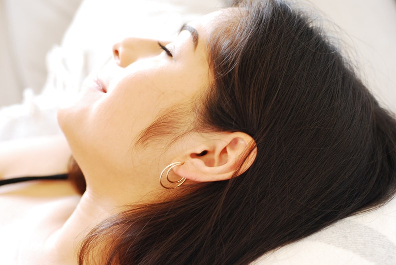May include: A close-up of a woman's face with her eyes closed. She is lying down with her head resting on a white pillow. Her hair is long and dark, and she is wearing a pair of small, silver hoop earrings.