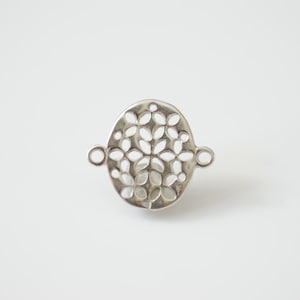 May include: Silver-toned metal connector with a circular, openwork design featuring a floral pattern. The connector has two loops on either side for attaching jewelry.