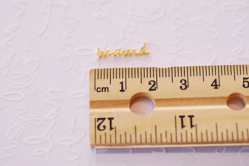 May include: A gold-colored metal word charm that reads "brand" is placed on a wooden ruler with centimeter markings. The ruler is marked from 1 to 5 centimeters.
