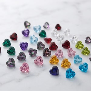 Pode incluir: Uma coleção de pequenas pedras preciosas facetadas em forma de coração numa variedade de cores vibrantes, incluindo vermelho, azul, verde, amarelo, rosa, roxo e transparente. Cada pedra apresenta um corte preciso que reflete a luz.