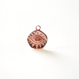 Puede incluir: Un amuleto de oro rosa con un dise&ntilde;o de ojo. El ojo est&aacute; rodeado de un patr&oacute;n de estrella. El amuleto tiene un bucle en la parte superior para sujetarlo a un collar o pulsera.