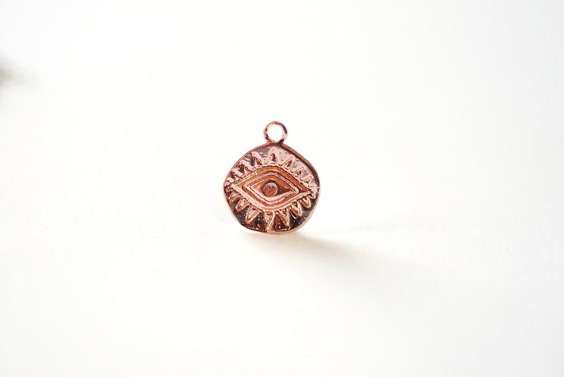 Puede incluir: Un amuleto de oro rosa con un dise&ntilde;o de ojo. El ojo est&aacute; rodeado de un patr&oacute;n de estrella. El amuleto tiene un bucle en la parte superior para sujetarlo a un collar o pulsera.