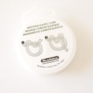 May include: White round container with instructions for knotting elastic cord. The container has black text that reads "Knotting Elastic Cord", "Nouage Cordon Elastique", "Anudando El Cordon Elastico", "Beadalon", "www.beadalon.com", "Beadalon - Valley Twp. PA 19320".