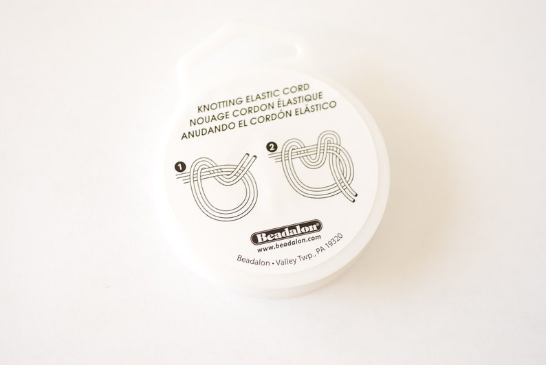 May include: White round container with instructions for knotting elastic cord. The container has black text that reads "Knotting Elastic Cord", "Nouage Cordon Elastique", "Anudando El Cordon Elastico", "Beadalon", "www.beadalon.com", "Beadalon - Valley Twp. PA 19320".