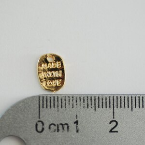 May include: Gold-coloured metal charm with the words "Made with Love" engraved on it. The charm is approximately 1 cm in diameter.