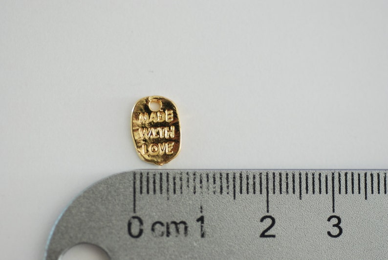 May include: Gold-coloured metal charm with the words "Made with Love" engraved on it. The charm is approximately 1 cm in diameter.