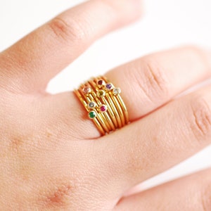 May include: A stack of gold rings with small colorful gemstones. The rings are worn on a person's finger.