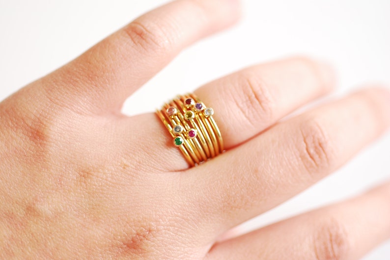 May include: A stack of gold rings with small colorful gemstones. The rings are worn on a person's finger.
