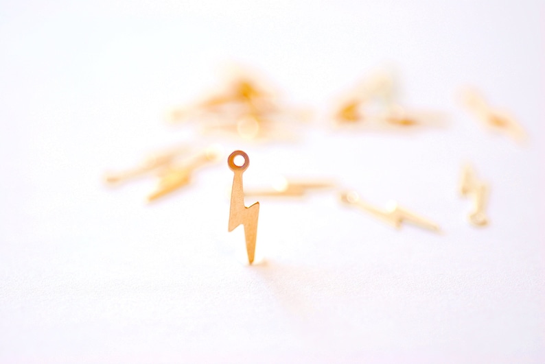 May include: Gold-colored metal lightning bolt charm with a jump ring. The charm is on a white surface.