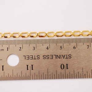 Puede incluir: Una cadena de color dorado con eslabones cuadrados, medida con una regla. La cadena mide aproximadamente 18 cm de largo.