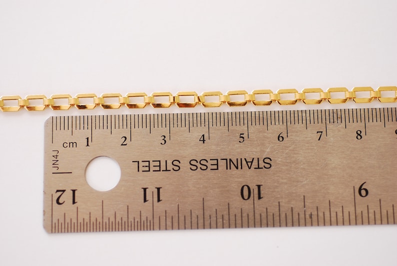 Puede incluir: Una cadena de color dorado con eslabones cuadrados, medida con una regla. La cadena mide aproximadamente 18 cm de largo.
